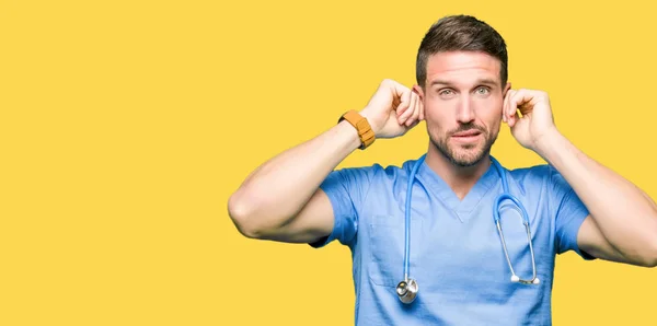 Handsome doctor wearing medical uniform over isolated background covering ears with fingers with annoyed expression for the noise of loud music. Deaf concept.