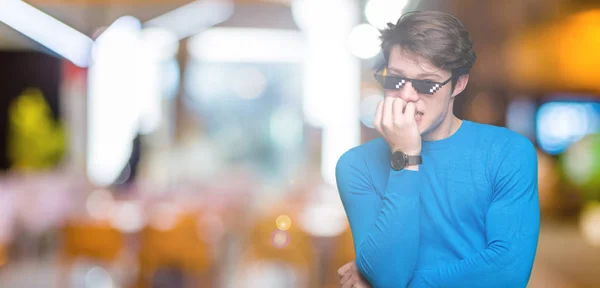 Young man wearing funny thug life glasses over isolated background looking stressed and nervous with hands on mouth biting nails. Anxiety problem.