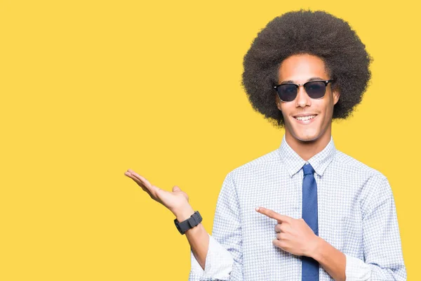 Young african american business man with afro hair wearing sunglasses amazed and smiling to the camera while presenting with hand and pointing with finger.