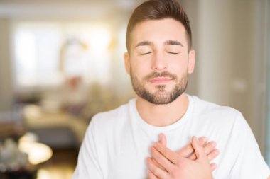 Yakışıklı delikanlı evde rahat beyaz t-shirt giyen kapalı gözler ile göğüs ellerinde ve yüzünde minnettar jest ile gülümseyen. Sağlık kavramı.