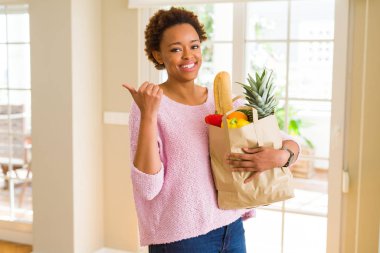 Kağıt torba işaret ve başparmak kadar mutlu yüz gülümseyen tarafı ile gösterilen taze yiyecek dolu tutan genç Afro-Amerikan kadın