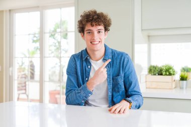 Genç yakışıklı adam giyiyor rahat denim ceket evde neşeli bir gülümseme ile el ve parmak kadar yan yüzünde mutlu ve doğal ifade ile işaret eden yüz
