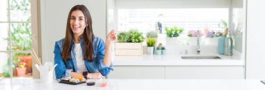 Kameraya bakarak yan el ve parmak ile işaret eden güzel genç kadın, yüzünde kocaman bir gülümseme ile teslim suşi yemek geniş açı resim.