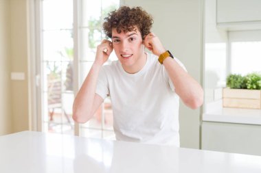 Evde beyaz t-shirt giyen genç yakışıklı adam yüksek sesle müzik gürültü rahatsız ifade parmaklarıyla kulaklarıyla kapsayan. Sağır kavramı.