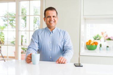 Orta yaş iş adamı ayakta ve diş gösterilen kendine güvenen bir gülümsemeyle gülümseyerek mutlu bir yüz ile sabahları evde kahve içme