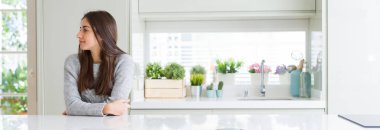 Güzel genç kadın evde beyaz masaya oturan geniş açı resim emin gülümseme ile doğal yüz pozuyla profil rahatla tarafına, seyir.