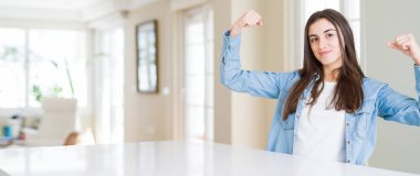 Geniş açı resim güzel genç kadın evde beyaz masaya gösteren kol kasları gururlu gülümseyen oturuyordu. Fitness kavramı.