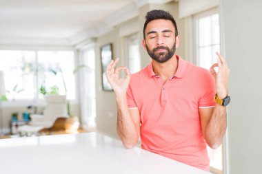 Evde rahat t-shirt giyen İspanyol yakışıklı rahatla ve gülümseyen gözlerle kapalı parmak jestle meditasyon yapıyor. Yoga kavramı.