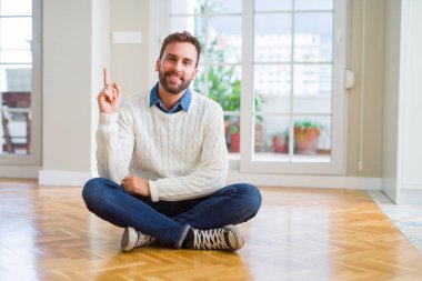 Yakışıklı adam yüzünde kocaman bir gülümseme ile yere evde oturmak rahat kazak giyen kameraya bakarak yan el ve parmak ile işaret.