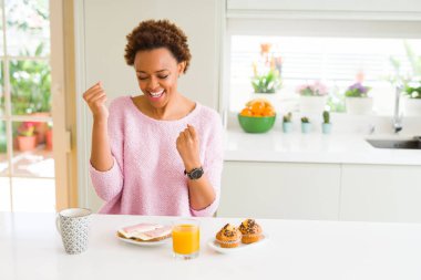 Gülümseyen ve başarı için çığlık evde çok mutlu ve heyecanlı kaldırdı, silah ile kazanan jest yapıyor sabah breaksfast yiyen genç Afro-Amerikan kadın. Kutlama kavramı.