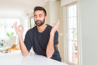Evde rahat kazak giyen yakışıklı İspanyol adam sakin ol ve gülümseyen gözlerle kapalı parmak jestle meditasyon yapıyor. Yoga kavramı.