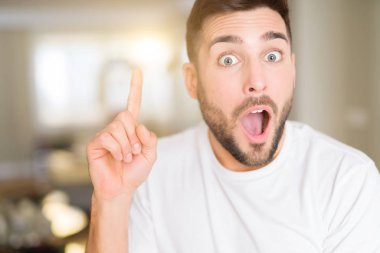 Evde rahat beyaz t-shirt giyen genç yakışıklı adam başarılı fikirle parmak işaret. Çıktıktan ve mutlu. Bir numara.