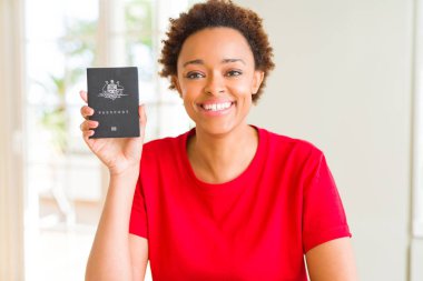 Avustralya pasaportu ile ayakta ve diş gösterilen kendine güvenen bir gülümsemeyle gülümseyerek mutlu bir yüz tutan genç Afro-Amerikan kadın