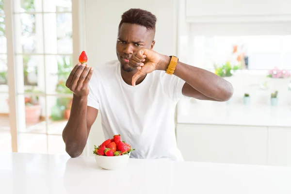African american man eating fresh healthy strawberries with angry face, negative sign showing dislike with thumbs down, rejection concept