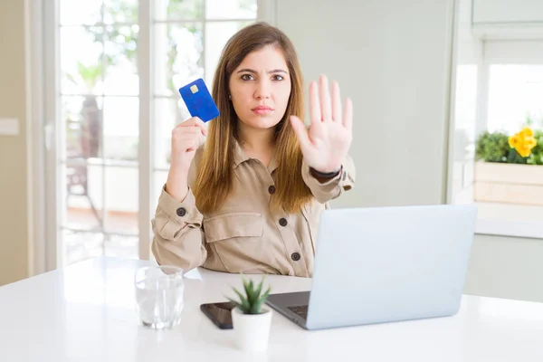 Beautiful young woman shopping online using laptop and credit card with open hand doing stop sign with serious and confident expression, defense gesture