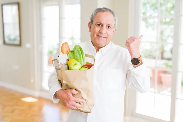 Handsome senior man holding a paper bag of fresh groceries from the supermarket pointing and showing with thumb up to the side with happy face smiling