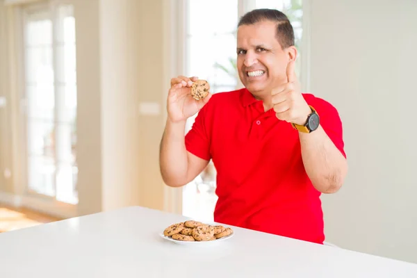 Middle age man eating chocolate chips cookies at home happy with big smile doing ok sign, thumb up with fingers, excellent sign