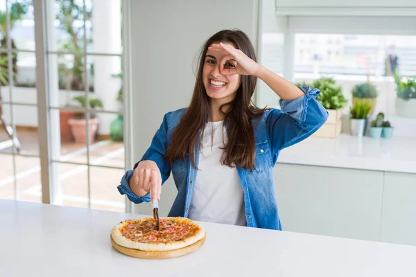 Beautiful young woman cutting a tasty pizza slice using a cutter with happy face smiling doing ok sign with hand on eye looking through fingers