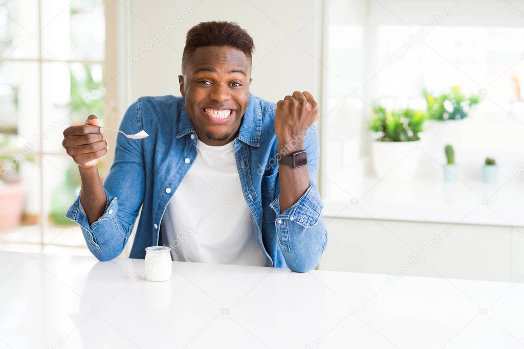 African american man eating healthy natural yogurt with a spoon screaming proud and celebrating victory and success very excited, cheering emotion