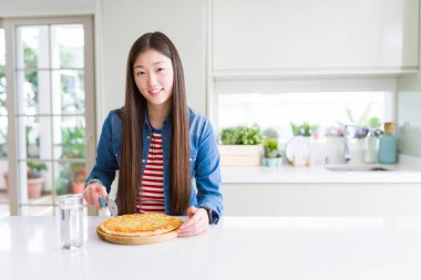 Yemek ve chesse ayakta ve diş gösterilen kendine güvenen bir gülümsemeyle gülümseyerek mutlu bir yüz ile pizza kesme güzel Asyalı kadın