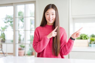 Gülümseyen ve iki ile işaret kamera bakıyor beyaz masa üzerinde pembe kazak giyen güzel Asyalı kadın eller ve yan parmak.