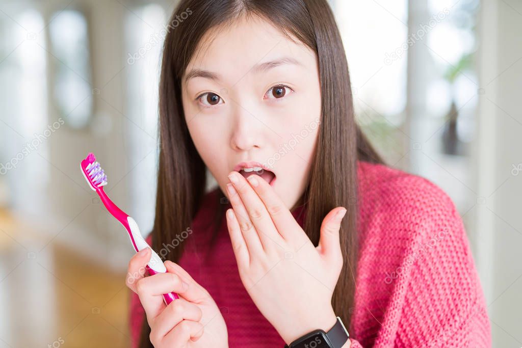 Beautiful Asian woman holding toothbrush cover mouth with hand shocked with shame for mistake, expression of fear, scared in silence, secret concept