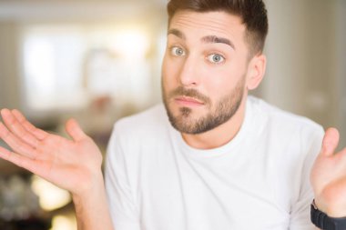 Genç yakışıklı giyiyor rahat beyaz t-shirt ev clueless ve şaşkın bir ifadeyle kollar ve eller kaldırdı, adam. Şüphe kavramı.