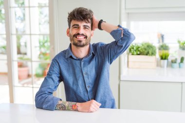 El hareketi ile kendine güvenen dokunaklı saç gülümseyerek, çekici poz beyaz masaya oturan rahat gömlek giymiş genç adam