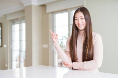 Kameraya bakarak yan el ve parmak ile işaret yüzünde kocaman bir gülümseme ile beyaz masada rahat kazak giyen güzel Asyalı kadın.