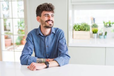 Genç giyiyor rahat gömlek beyaz uzak tarafına gülümseme ile yüzünde, doğal ifade seyir tablo oturan adam. Kendine güvenen gülüyor.