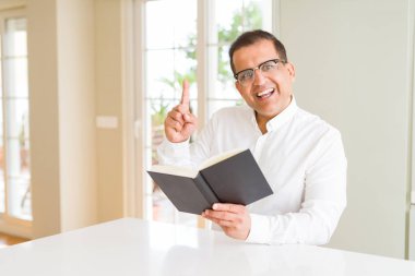 Orta yaş erkek evde kitap okurken bir fikirle sürpriz gözlük ya da gülen yüz, bir numara ile işaret parmağı soru