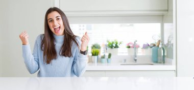 Güzel genç kadın evde beyaz masaya oturan geniş açı resim şaşırmış ve başarı için hayran kaldırdı Kollarını açarak kutluyor ve gözleri açık. Kazanan kavramı.