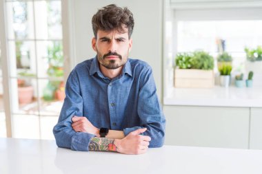 Beyaz masa şüpheci ve sinir, onaylamayan ifade yüz çapraz kolları üzerinde oturan rahat gömlek giymiş genç adam. Negatif kişi.