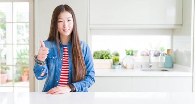 Beautiful Asian woman wearing denim jacket on white table doing happy thumbs up gesture with hand. Approving expression looking at the camera showing success. clipart