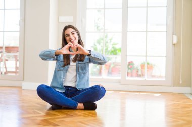 Kalp simgesi ve elleriyle şekil gösterilen aşık yere evde oturmak güzel genç kadın gülümseyerek. Romantik kavramı.