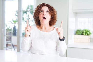 Evde beyaz kazak giyen güzel kadın kıdemli hayran ve yukarıya bakıyor ve parmakları ile işaret ve silah kaldırdı şaşırttı.