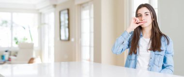 Ağız ve dudaklar evde beyaz masaya oturan güzel genç kadın geniş açı resim olarak ZIP parmak ile kapatın. Gizli ve sessiz, tabu söz