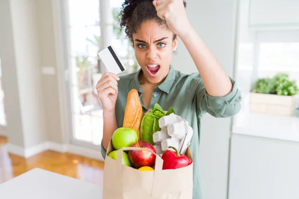 Young african american girl holding paper bag of groceries and credit card as payment annoyed and frustrated shouting with anger, crazy and yelling with raised hand, anger concept