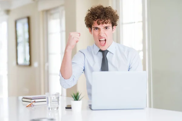 Young business man working with computer laptop at the office angry and mad raising fist frustrated and furious while shouting with anger. Rage and aggressive concept.
