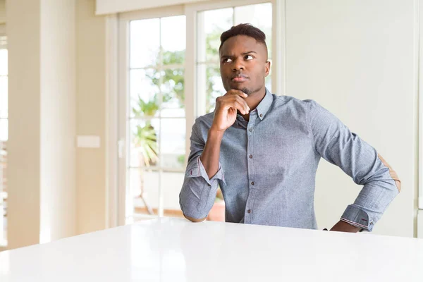 Handsome african american man on white table with hand on chin thinking about question, pensive expression. Smiling with thoughtful face. Doubt concept.