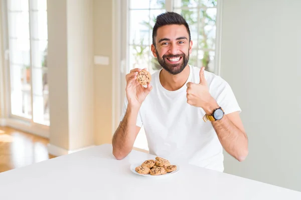 Handsome hispanic man eating chocolate chips cookies happy with big smile doing ok sign, thumb up with fingers, excellent sign