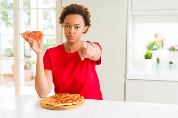 Young african american woman eating tasty peperoni pizza pointing with finger to the camera and to you, hand sign, positive and confident gesture from the front