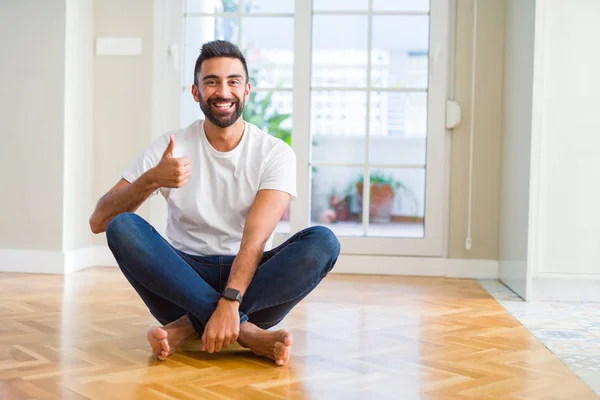 英俊的西班牙男子穿着休闲 T恤坐在地板上 在家里做快乐的大拇指向上手势用手 通过显示成功来批准镜头的表达 — 图库照片
