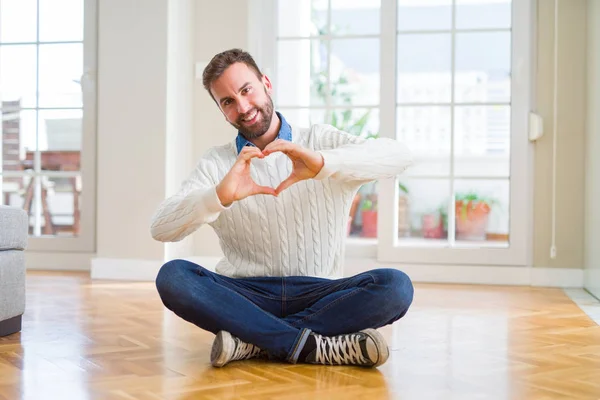 帅哥穿着休闲毛衣坐在家里的地板上 微笑着用双手展现心灵的象征和形状 浪漫的概念 — 图库照片