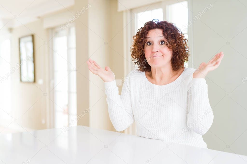 Beautiful senior woman wearing white sweater at home clueless and confused expression with arms and hands raised. Doubt concept.