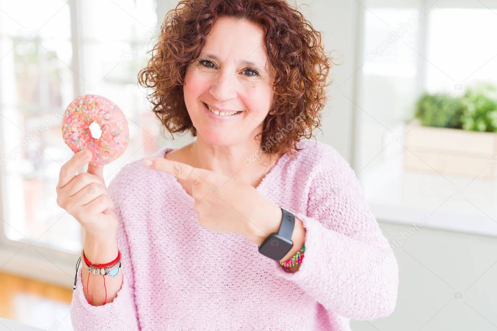 Senior woman eating pink sugar donut very happy pointing with hand and finger