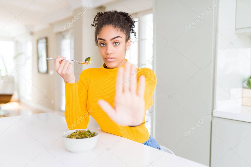Young african american woman eating healthy green vegatables with open hand doing stop sign with serious and confident expression, defense gesture