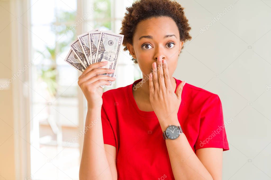 Young african american woman holding bank notes of fifty dollars cover mouth with hand shocked with shame for mistake, expression of fear, scared in silence, secret concept