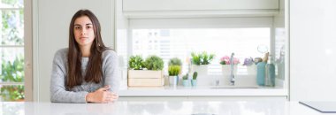 Geniş açı resim güzel genç kadının yüzünde ciddi bir ifade ile evde beyaz masaya oturuyor. Basit ve doğal kameraya bakıyor.