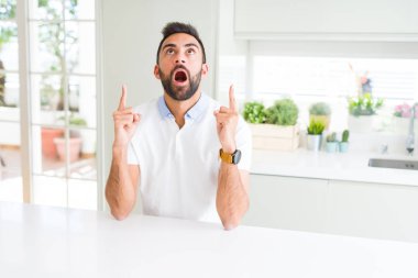 Casual beyaz t-shirt evde hayret ve yukarıya bakıp parmakları ile işaret şaşırttı ve silah kaldırdı İspanyol yakışıklı.